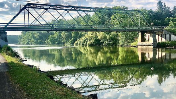 Still waters along a canal underneath a bridge with views of green forests in the background