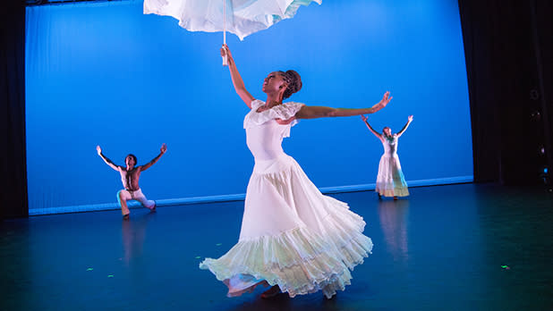 A trio of dancers dressed in white costumes perform on a stage with a blue background with the Alvin Ailey American Dance Theater