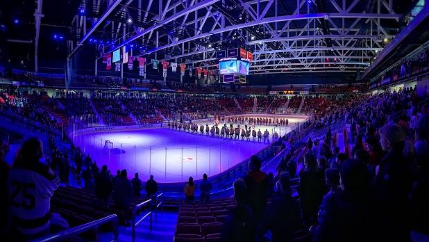 Audience watches hockey players gathered in a line under spotlights on the ice at the Lake Placid Olympic Center