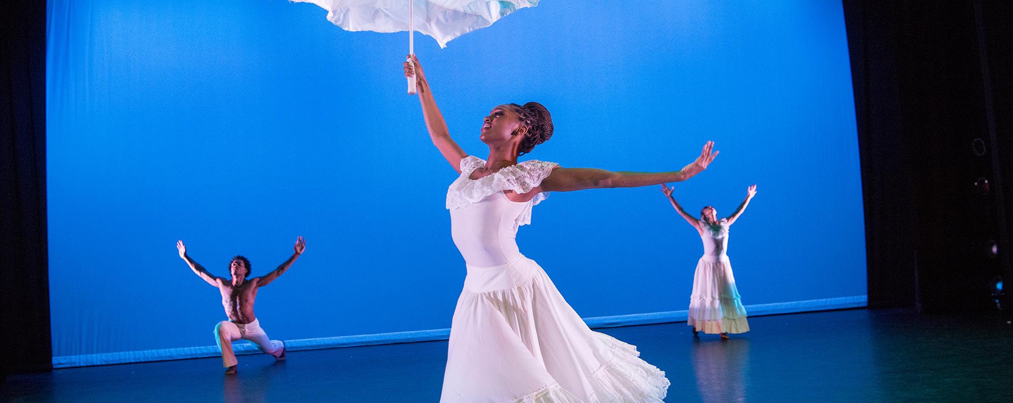 Two female and one male dancer dressed in white dance in front of a blue backdrop at the Alvin Ailey American Dance Theater