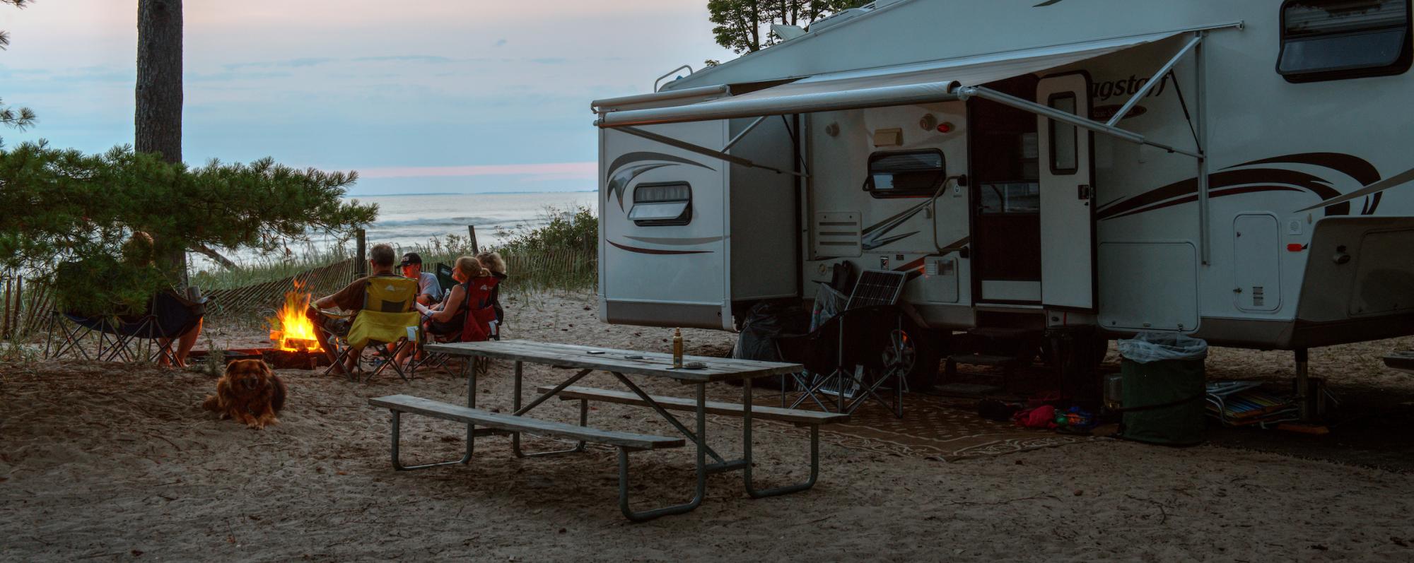 A family sits by a fire pit alongside their white camper at Southwick Beach State Park